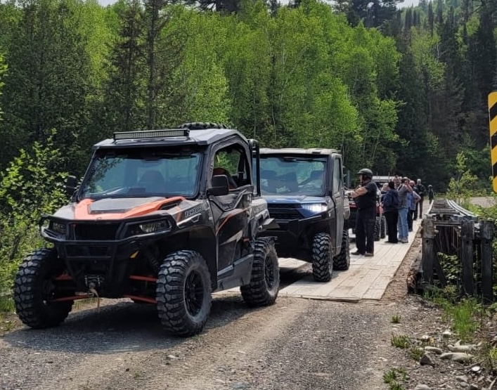 a line of UTVs parked on a narrow wooden bridge on a lush forest trail. Riders standing next to the UTVs discuss the route.