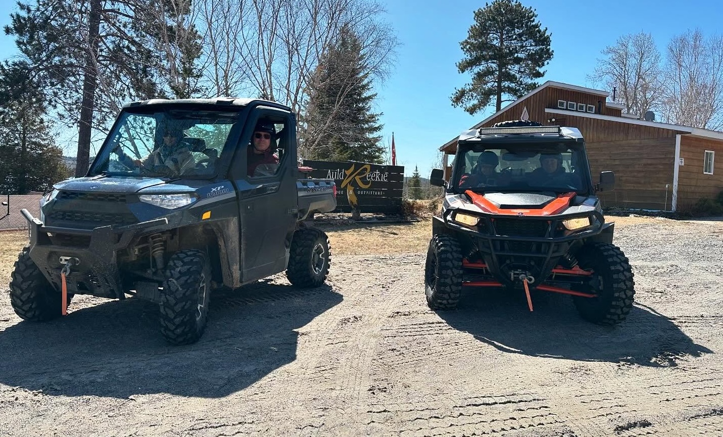 Riders look out of two UTVs parked in front of a wooden Auld Reekie Lodge sign on a summer day. There is forest behind them and the dusty trail around them is loaded with tire tracks.