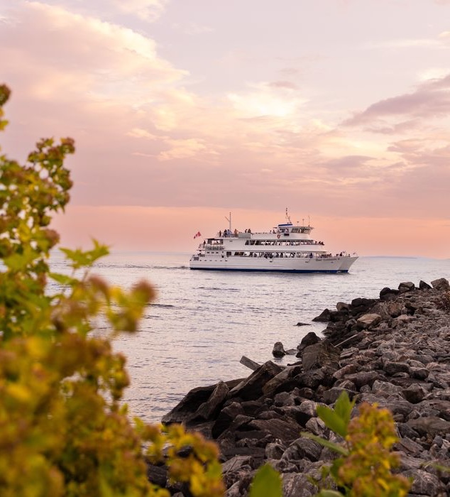 The Chief Commanda II sailing during a soft rose-coloured sunset on shining waters near a rocky forested bank. 