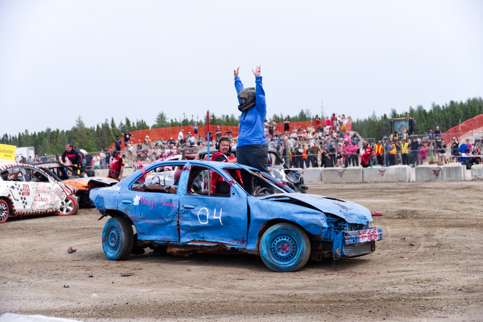 a driver in a helmet stands next to their smashed up derby car, hands raised above their head and cheering while the crowd in the stands looks on.