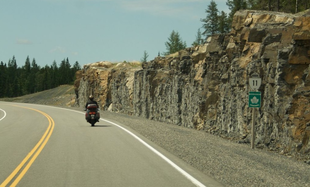 A motorcyclist rides along an empty stretch of highway with a "11" highway marker sign at the side of the road. Steep rock cliffs and forest line the side of the road.