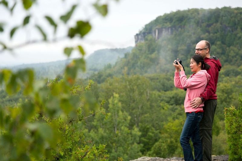 A man and woman stand close as the woman smiles and snaps a photo of the beautiful lush green forest and rocky cliffs that surround them. 