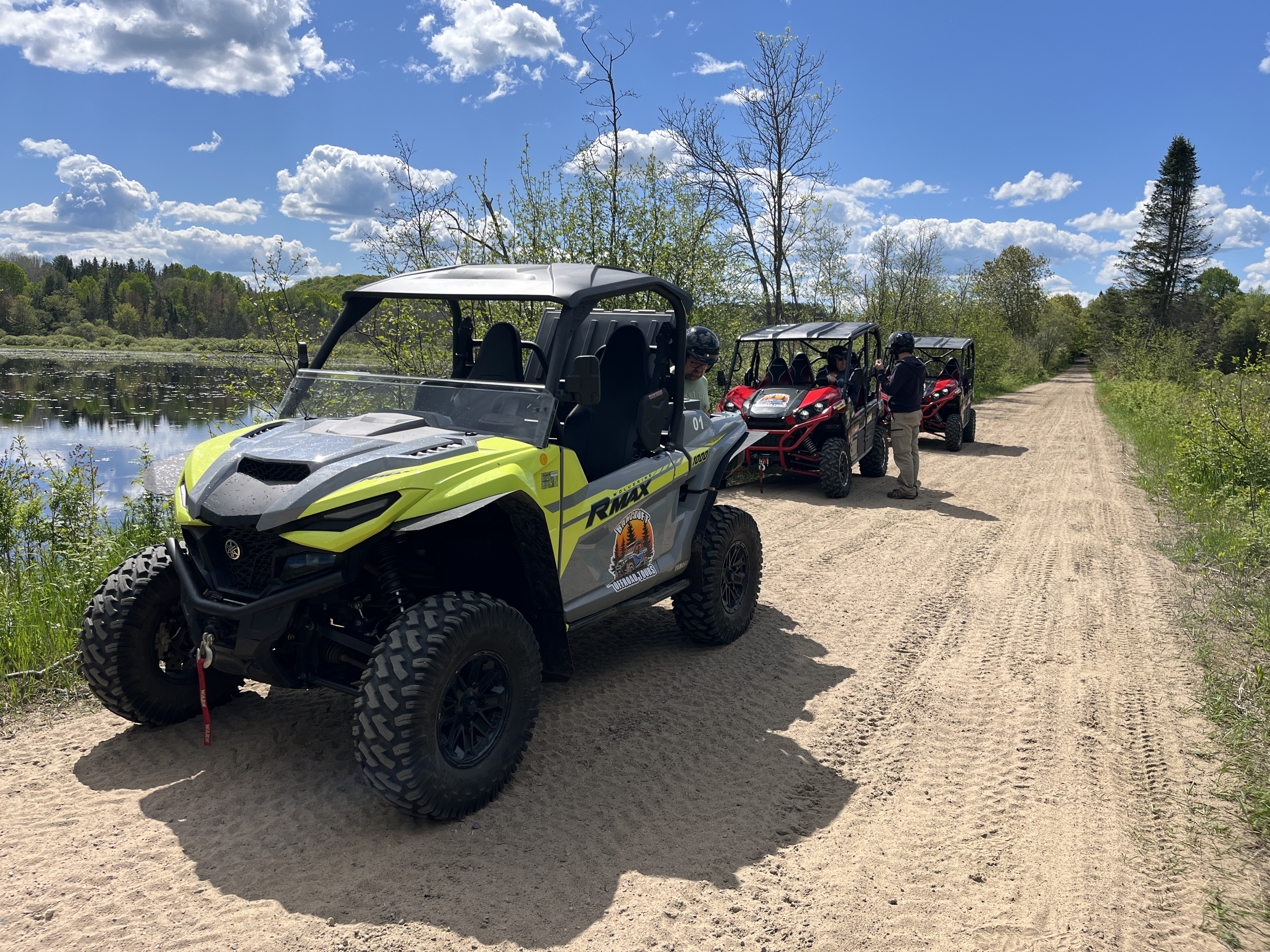 3 UTVs parked on a dusty trail along a lake, surrounded by green trees and grasses. One rider in a helmet talks to another. 