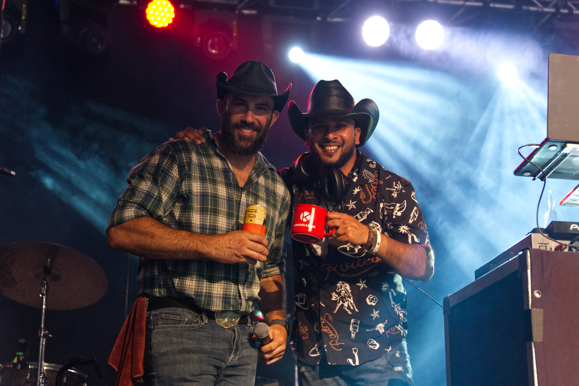 Two men wearing cowboy hats and plaid shirts smile as they hold up drinks. They are standing next to a DJ table while coloured spotlights stream down in the background.