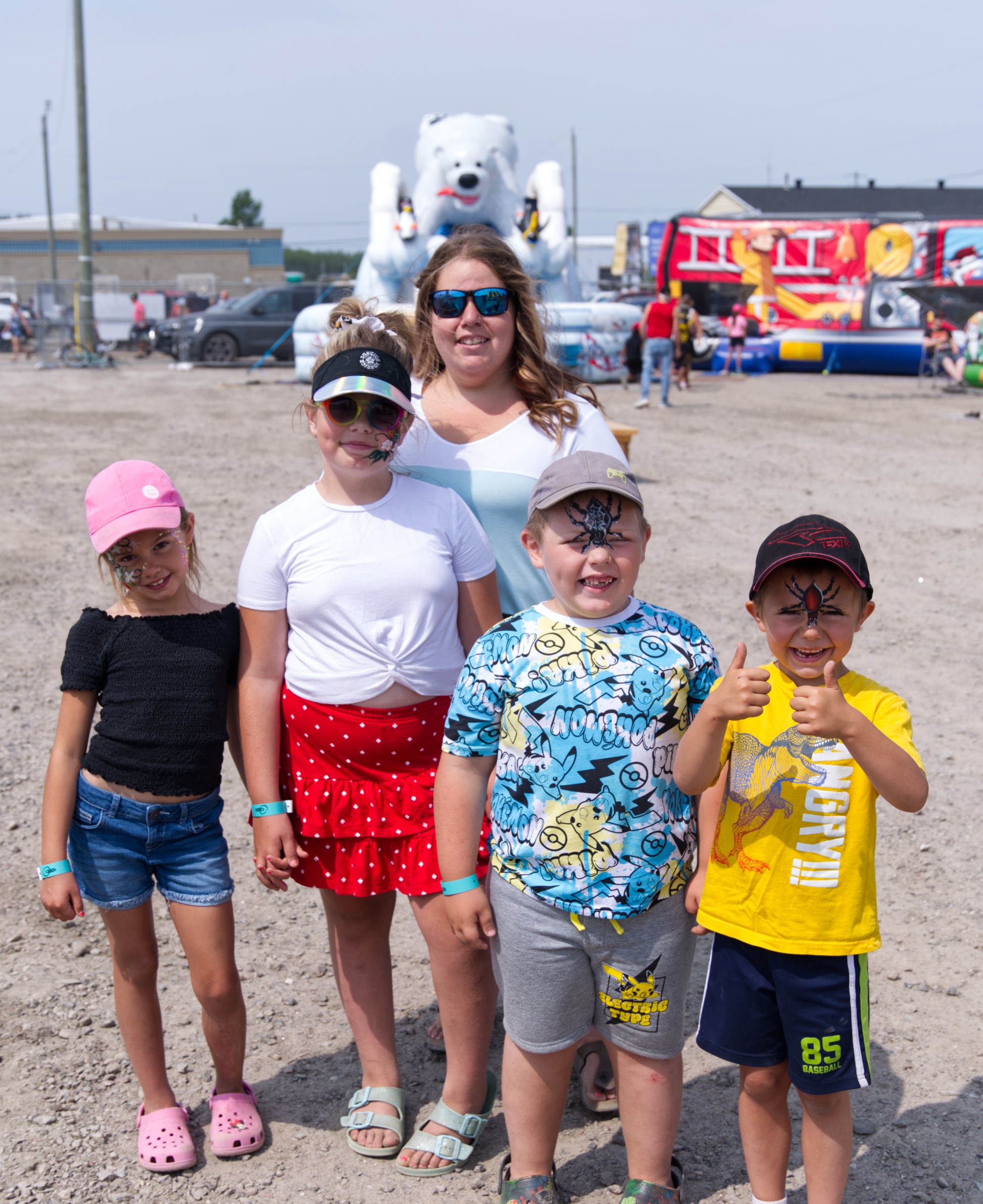 a row of kids with painted faces smile, one giving a thumbs-up and a big grin. Large inflatable bouncy castles are in the background.