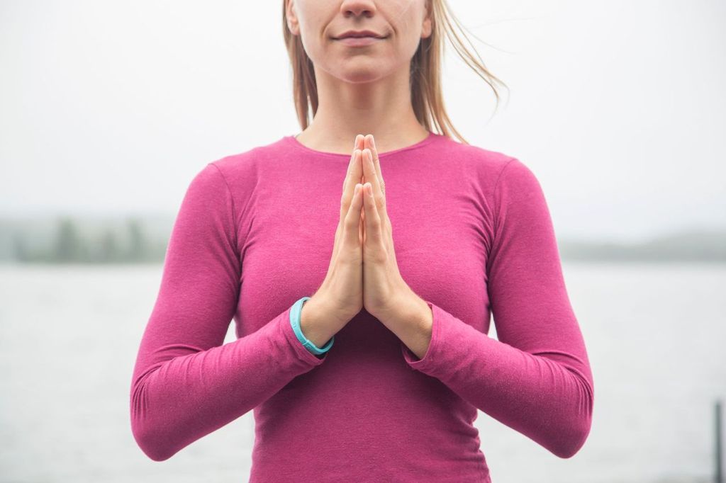 A woman smiles peacefully with her hands in Namaste on a breezy lakeside beach. 
