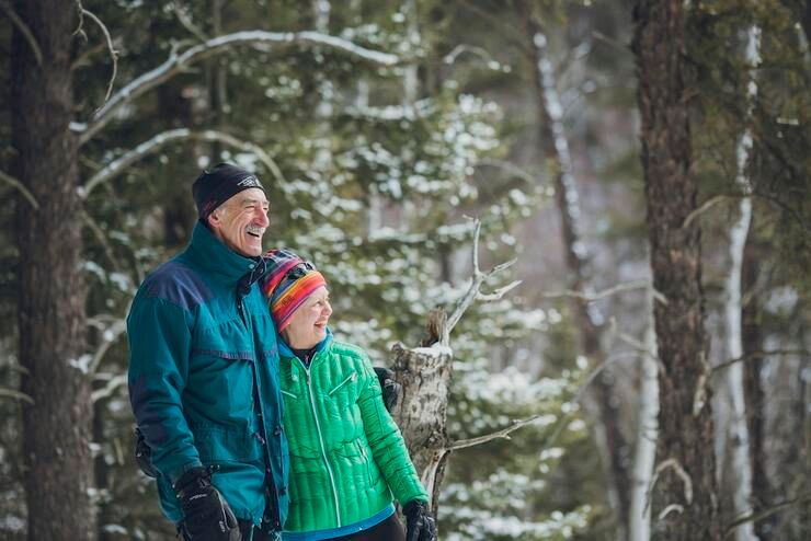 An older couple wearing winter gear smile and laugh as they look at the snow-covered forest around them. 