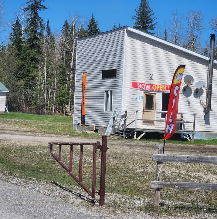 A simple white wooden building beyond an open metal gate, decked with a bright "Now Open!" sign and one advertising Wifi. A thick forest sits behind the property. 