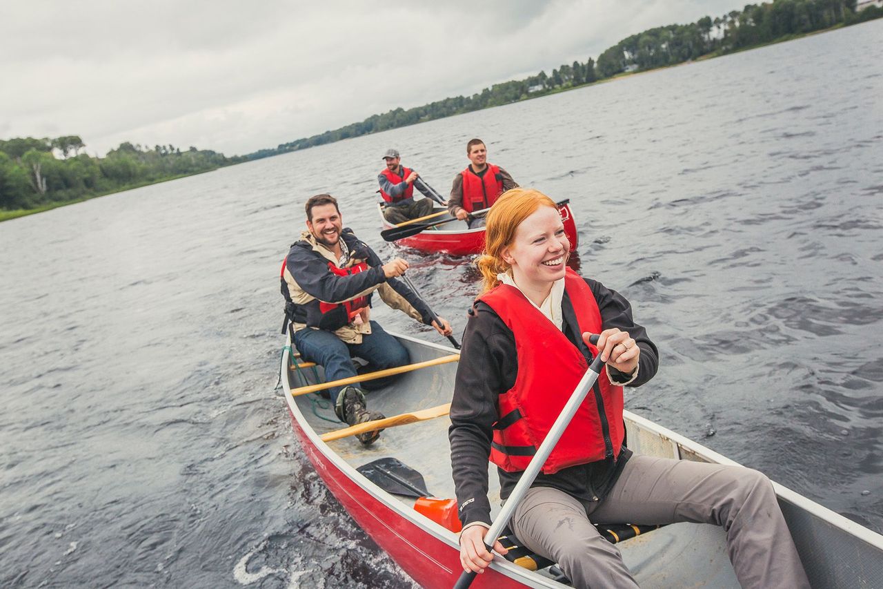 Four paddlers sharing two canoes all smile energetically as they paddle across a large lake on a cloudy summer day. Thick green forest sits on the horizon.