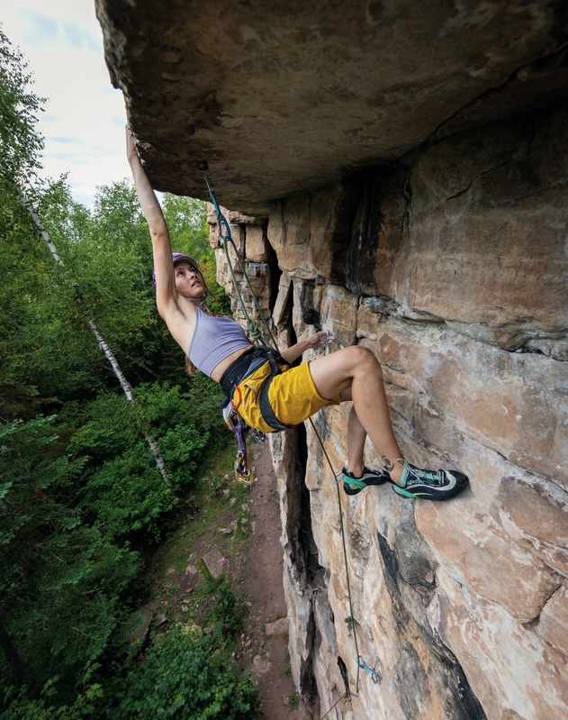 A woman wearing climbing gear looks determined and focused as she scales a vertical rock cliff with thick green forest below.
