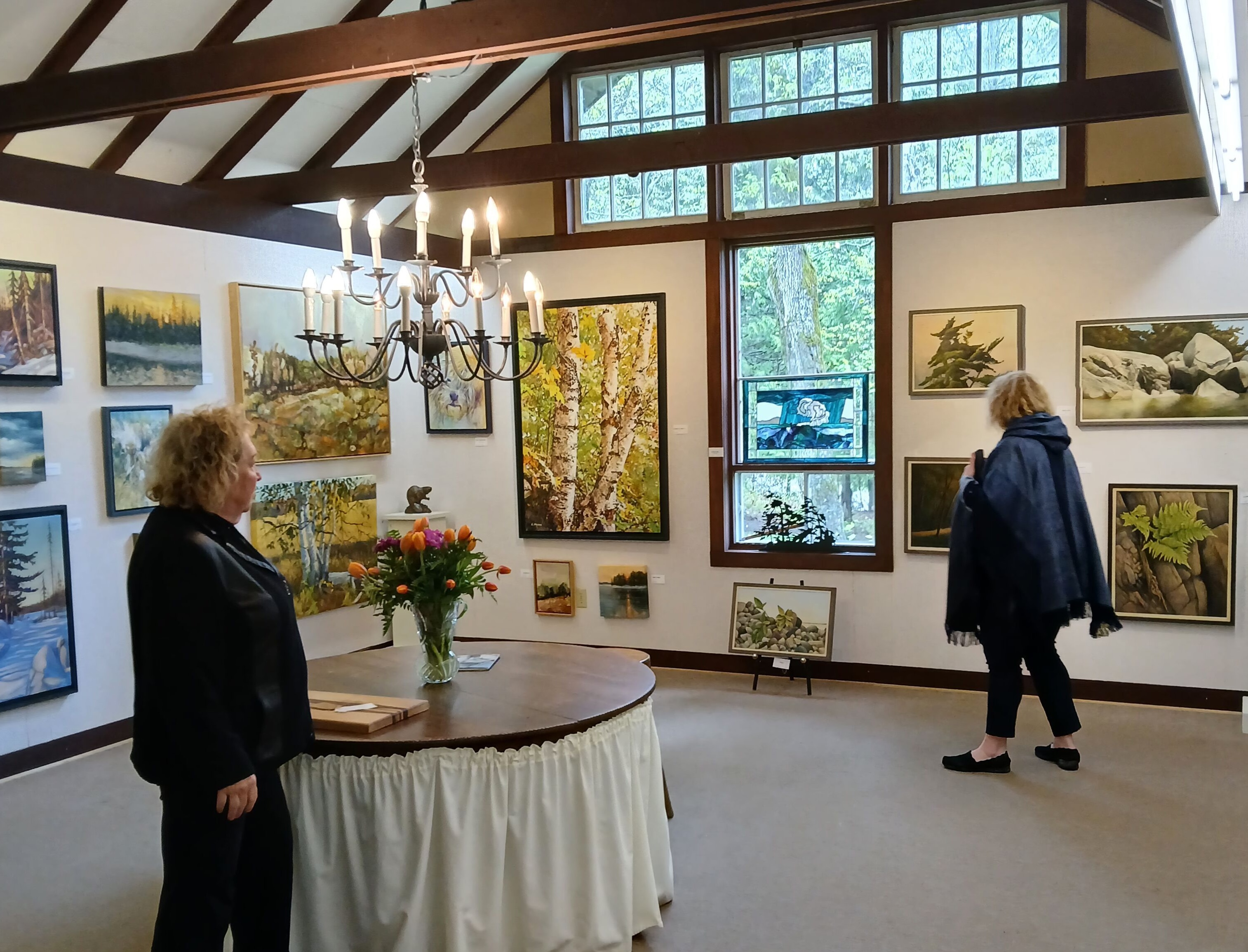 Two visitors peruse the series of beautiful paintings arranged on the Perivale Gallery walls, featuring nature motifs inspired by the Northern Ontario landscape.