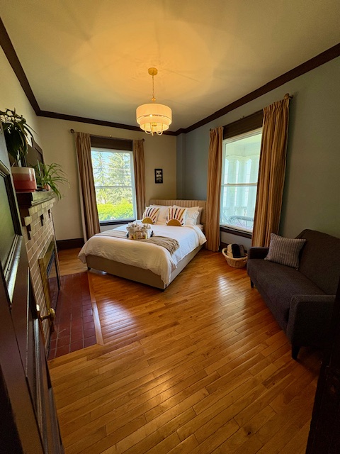 a comfortable bedroom with a luxurious bed, shining wood floors and tall antique windows. A glass chandelier adds soft lighting and green plants adorn a fireplace on one wall.