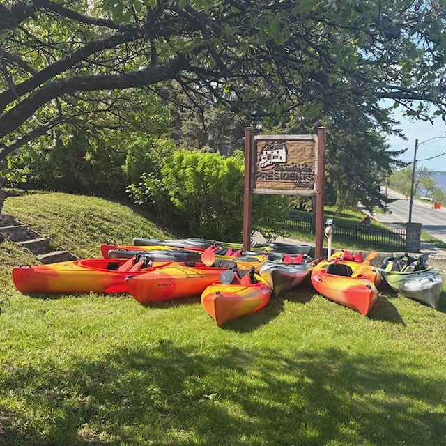 a row of kayaks laid in the lush green grass around a carved wooden sign that reads "The Presidents' Suites". Green shady trees hang overhead and a blue lake lies just beyond the kayaks.