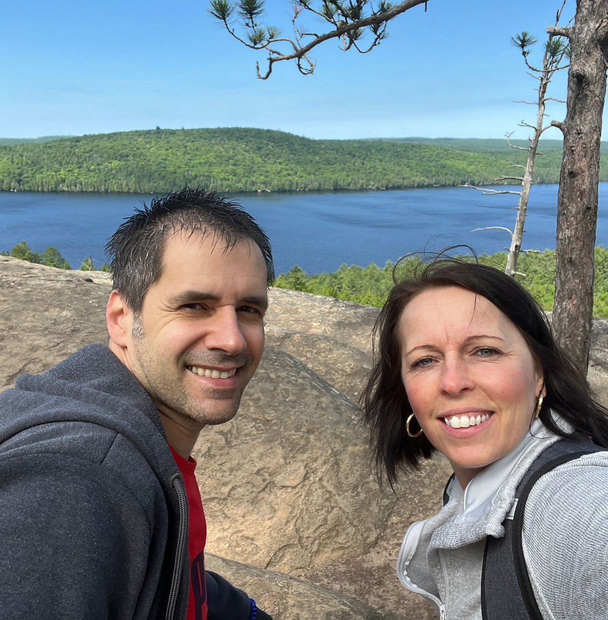 A man and woman with dark hair and casual clothing smile warmly in front of a blue lake with smooth rock banks and a green forested island on the horizon.
