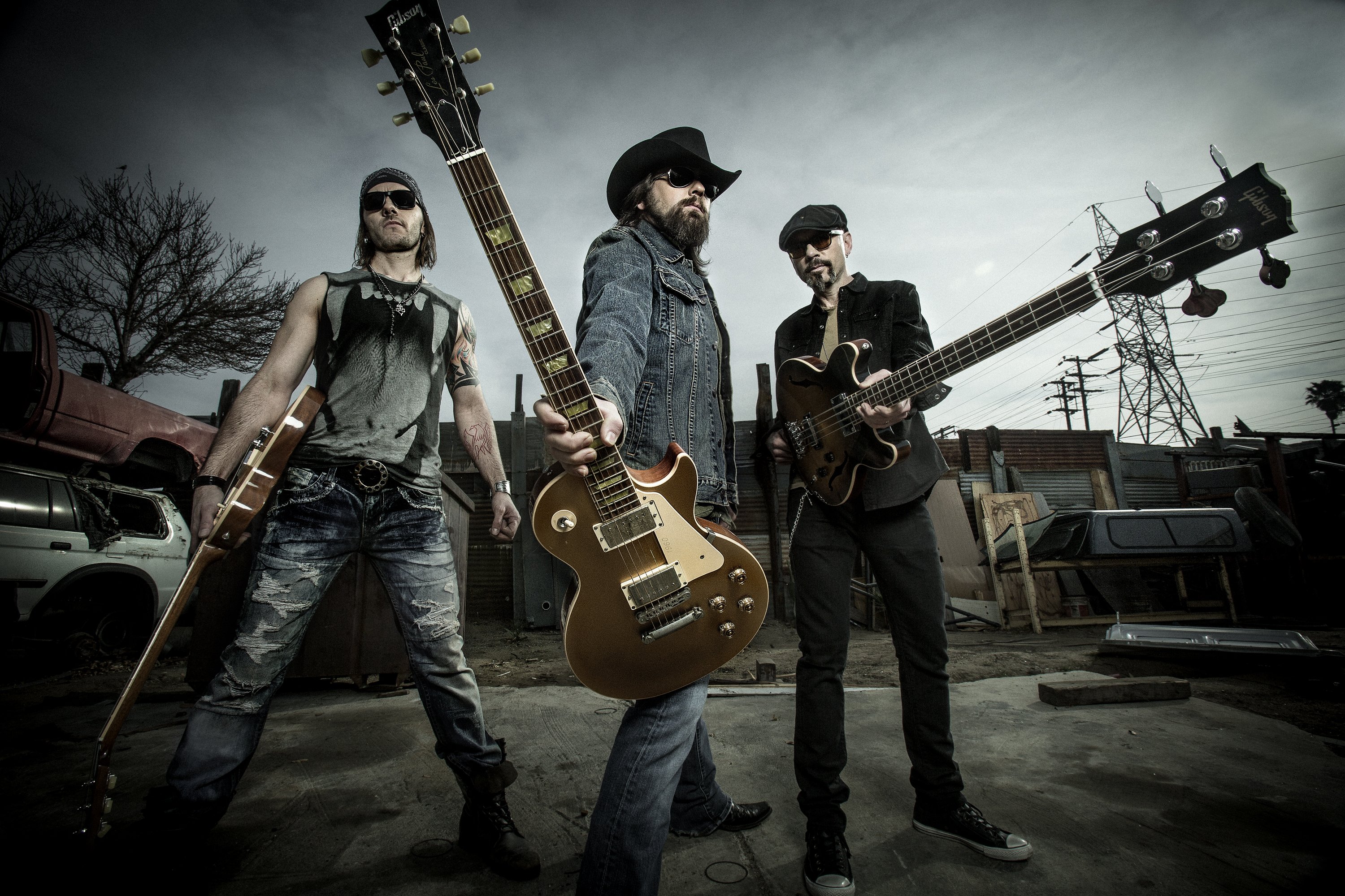 The Road Hammers; a band of 3 men wearing sunglasses, cowboy hats and denim pose dramatically with their guitars, looking down at the camera, in front of a moody grey outdoor background.