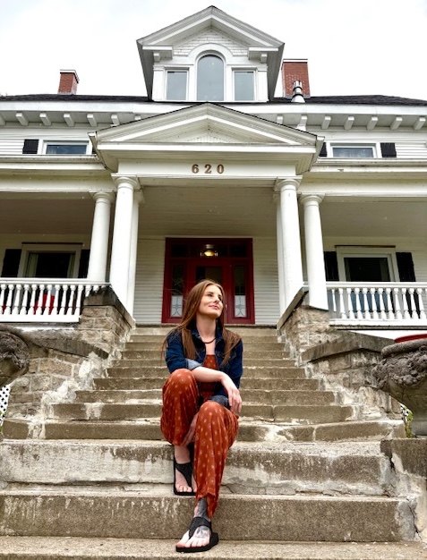 a smiling, stylishly dressed woman sits on the front steps of a heritage mansion in Haileybury, looking dreamily off into the distance.