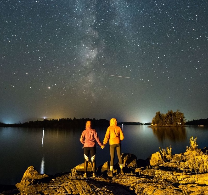 Two people hold hands as they look out into the night sky at a meterorite zipping through a milky sea of stars. A forest sits behind them and a shining lake stretches out in front of them. 