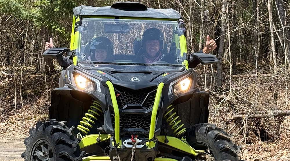 Two riders smile and flash thumbs-up out the windows of their UTV in the forest.
