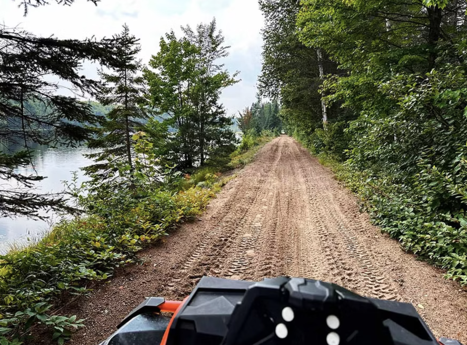 A dirt trail covered in tire tracks stretches out in front of an ATV through thick green forest next to a lake.