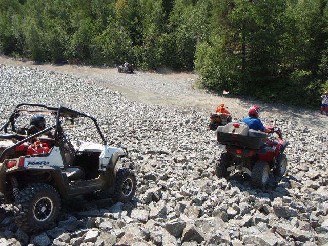 ATVers ride down a steep hill covered in large jagged rocks skirted by thick green forest.