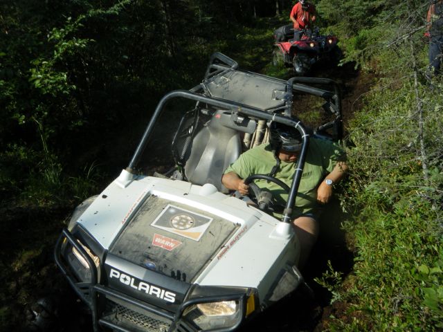 UTVs pass through dense green underbrush with water on the trail.
