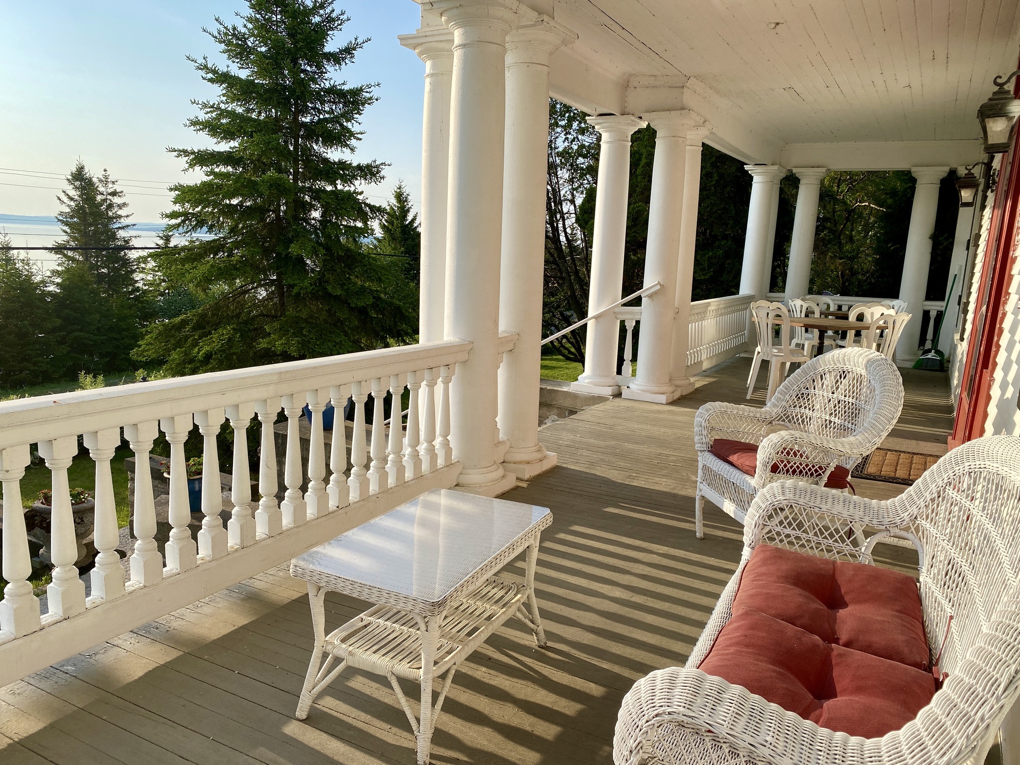 A set of comfortable white wicker chairs and sofa sit on a sunny covered porch at The President's Suites, looking out over a shining lake and trees. The stately white pillars and railing of the beautiful heritage building are in the background.