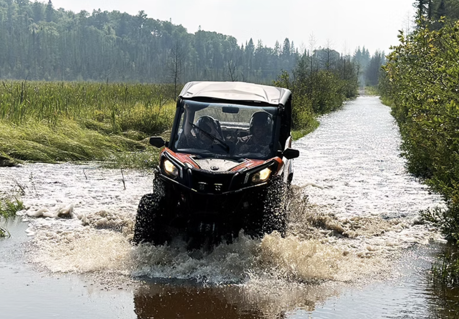 A UTV drives along a trail in a boggy clearing skirted by forest, the length of the trail entirely flooded with shining water on a sunny summer day. The UTV raises a large splash as it drives along.