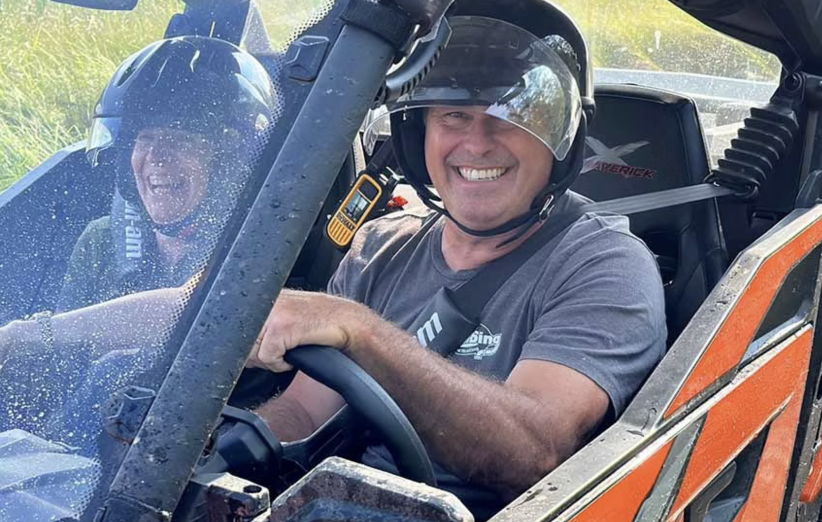 Two riders grinning and laughing as they drive a mud and water-spattered UTV.