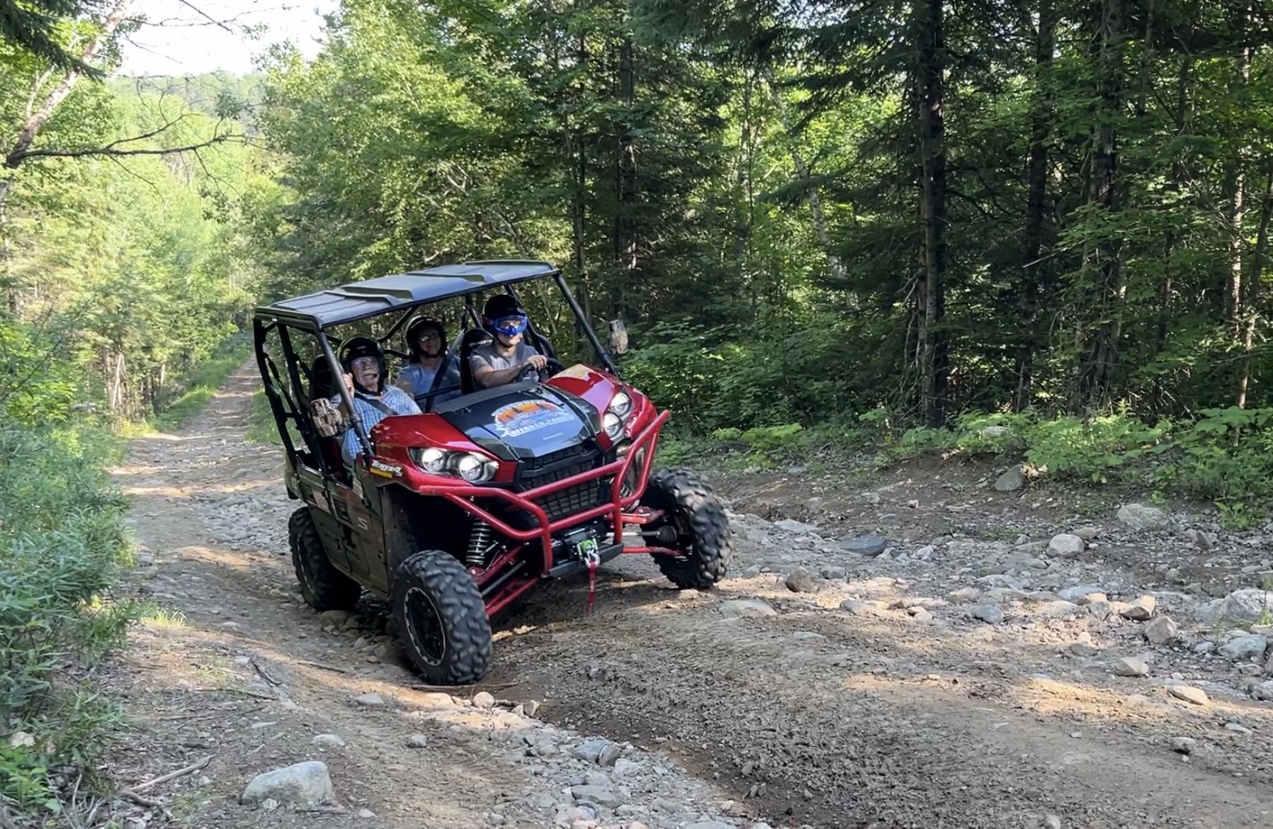 A UTV full of riders rolls down a rocky dirt trail surrounded by thick forest.