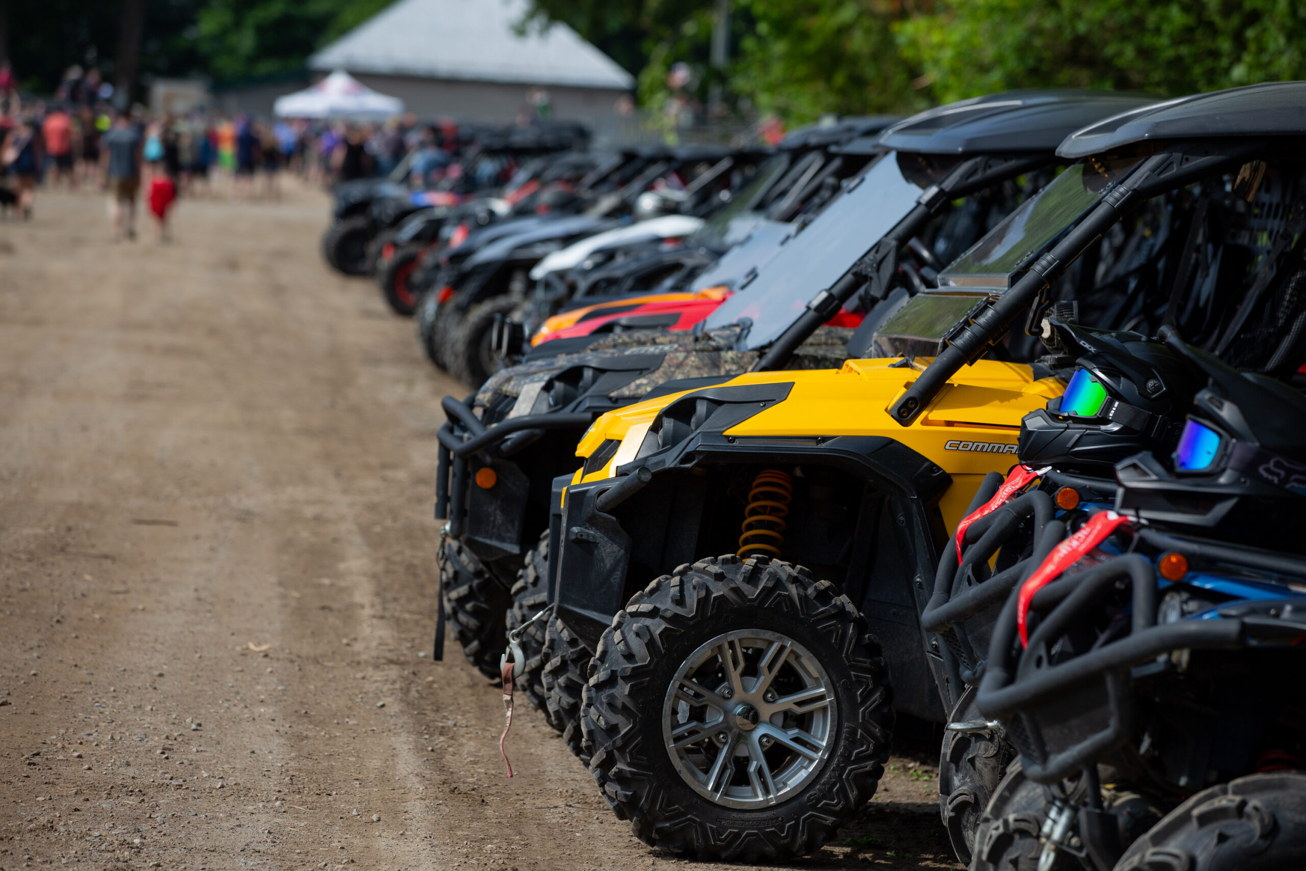a long line of parked ATVs of all kinds stretches into the distance, where eventgoers can be seen walking into the fairgrounds full of tents at the Rockin' the Trail summer event.