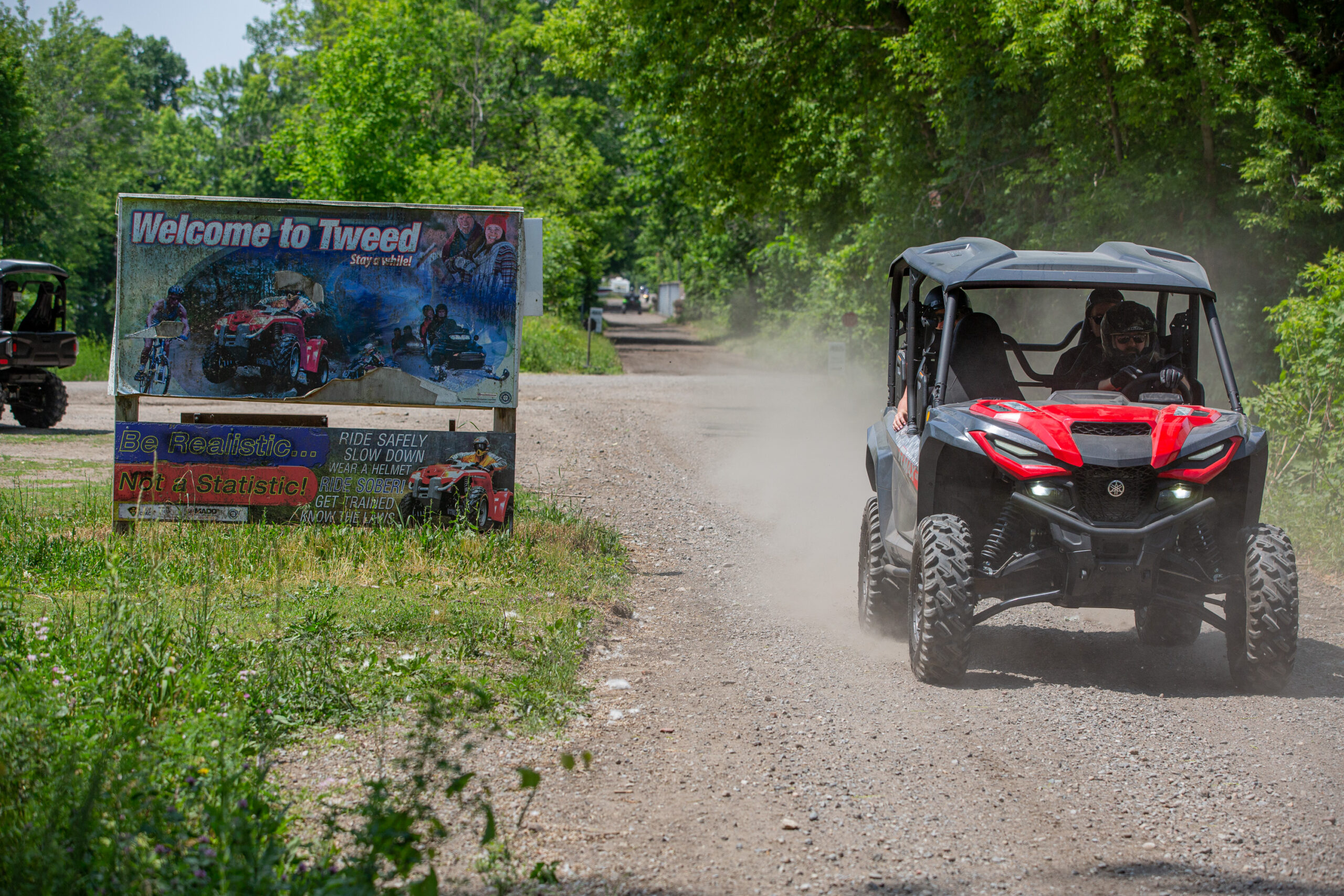 A UTV drives down a dusty gravel road surrounded by green forest past a small billboard that says "Welcome to Tweed".