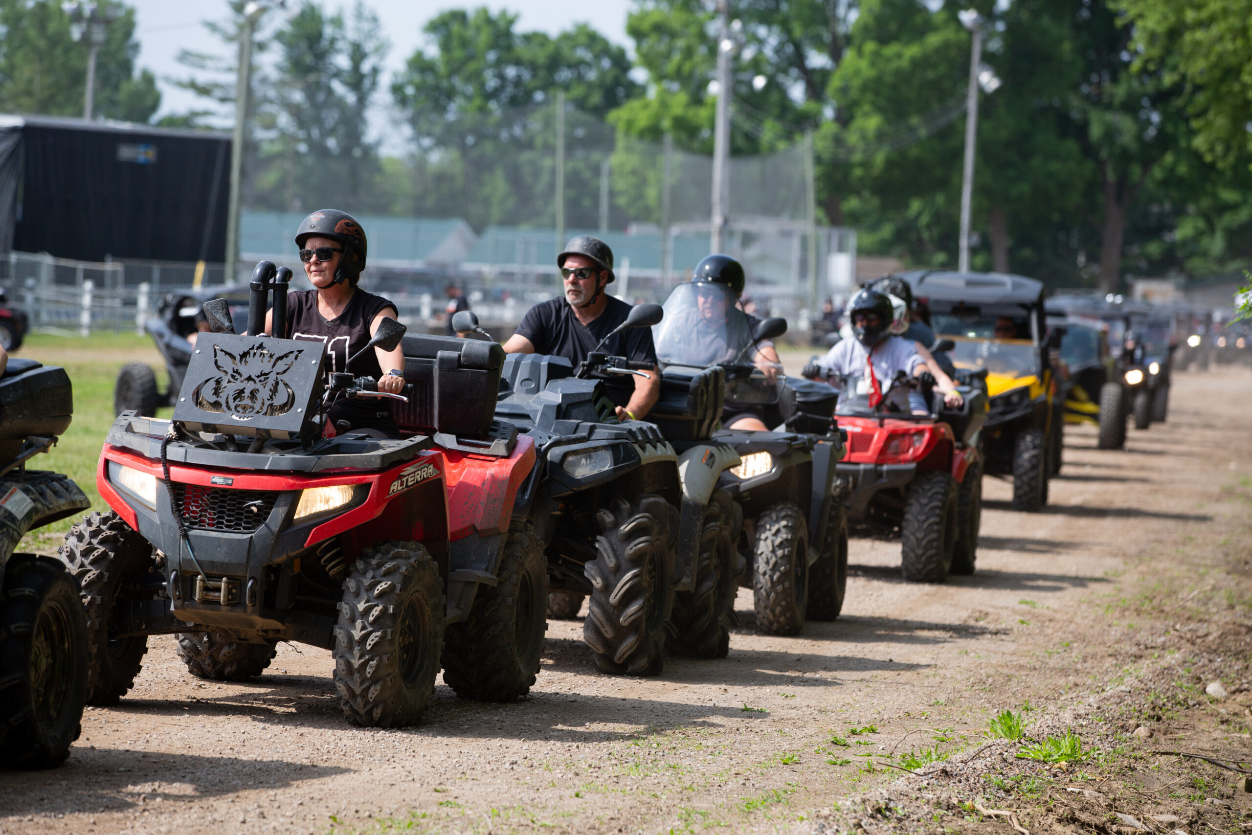 A long line of ATVs with smiling riders rolls down a gravel road at the Tweed Fairgrounds during the Rockin' the Trail event.