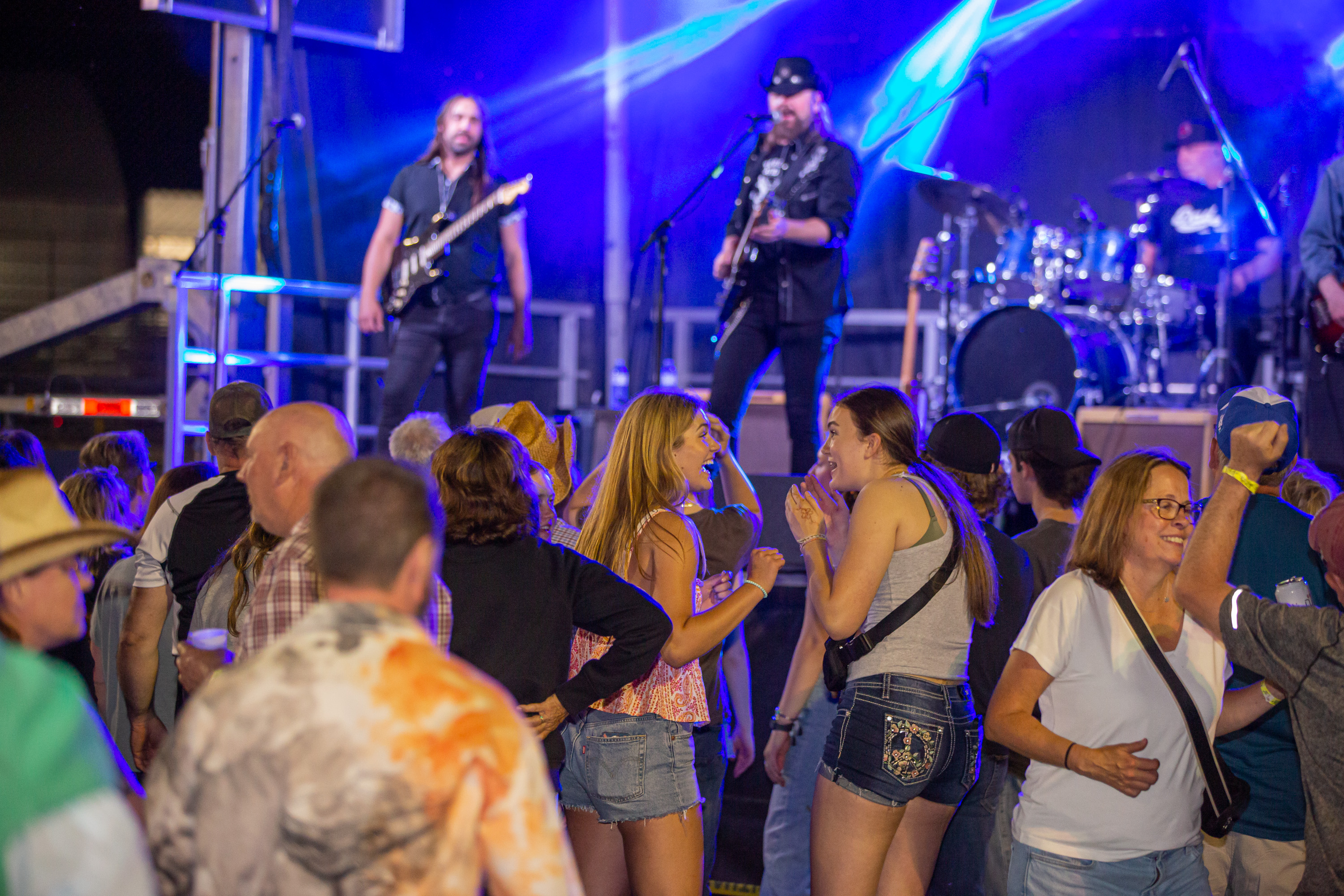 Two young women stand close together in a large crowd, talking and laughing with glee as they watch Jason McCoy and his band perform onstage at Rockin' the Trails 2025.