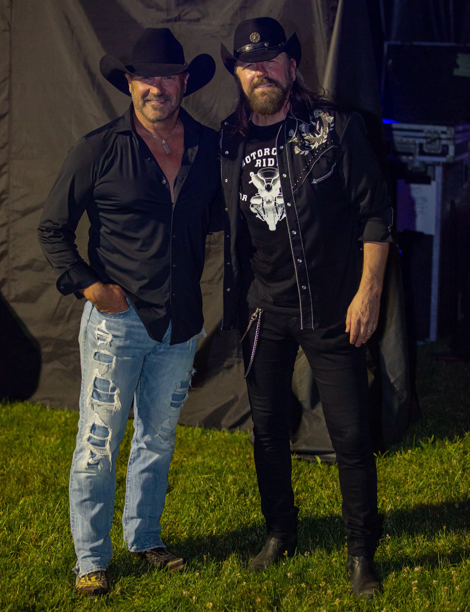 Country music artists Larry Berrio and Jason McCoy stand in green grass on a summer night. They are smiling for the camera backstage at Rockin' the Trails 2025.