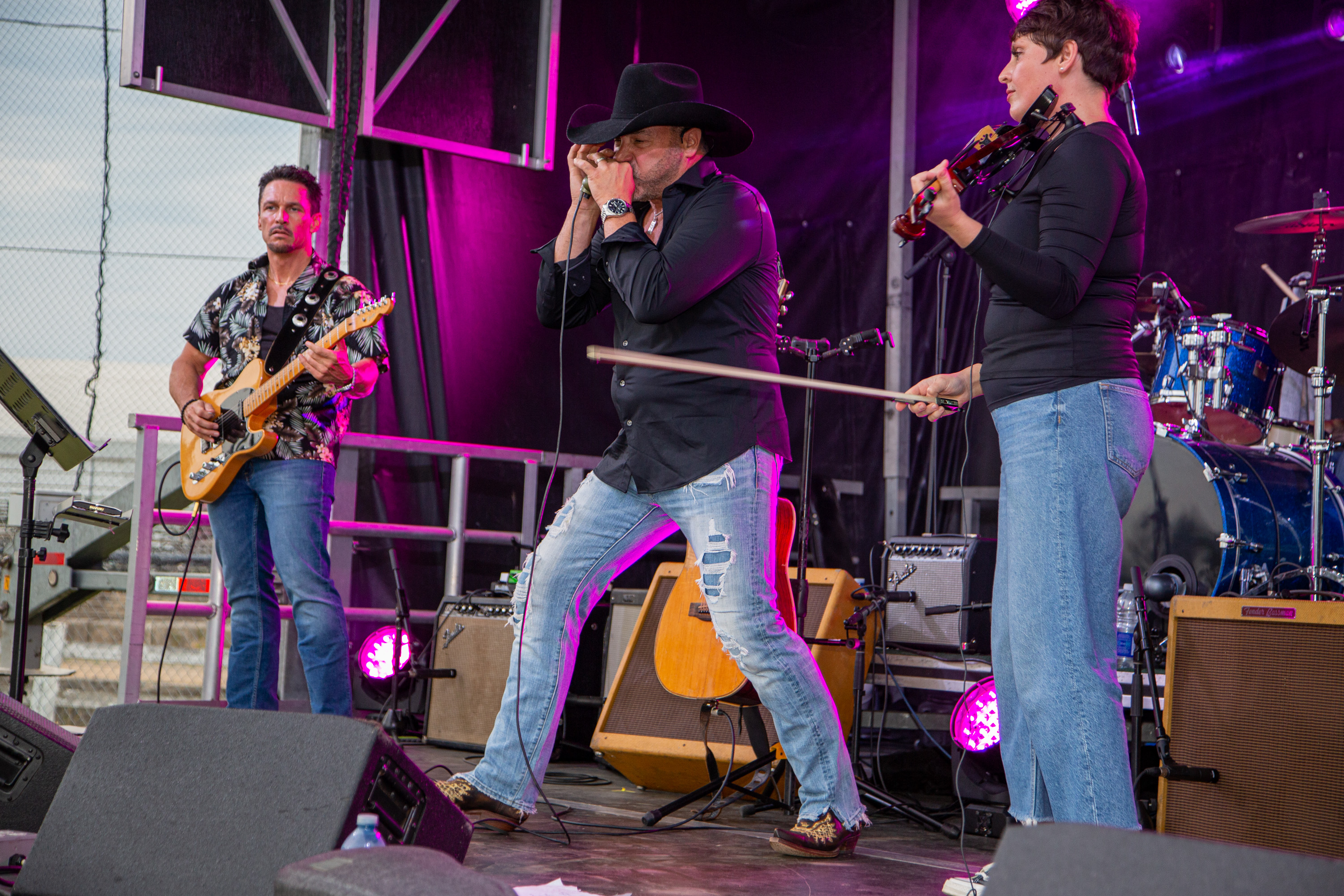 Country music artist Larry Berrio plays a harmonica on stage with his band members under purple spotlights.
