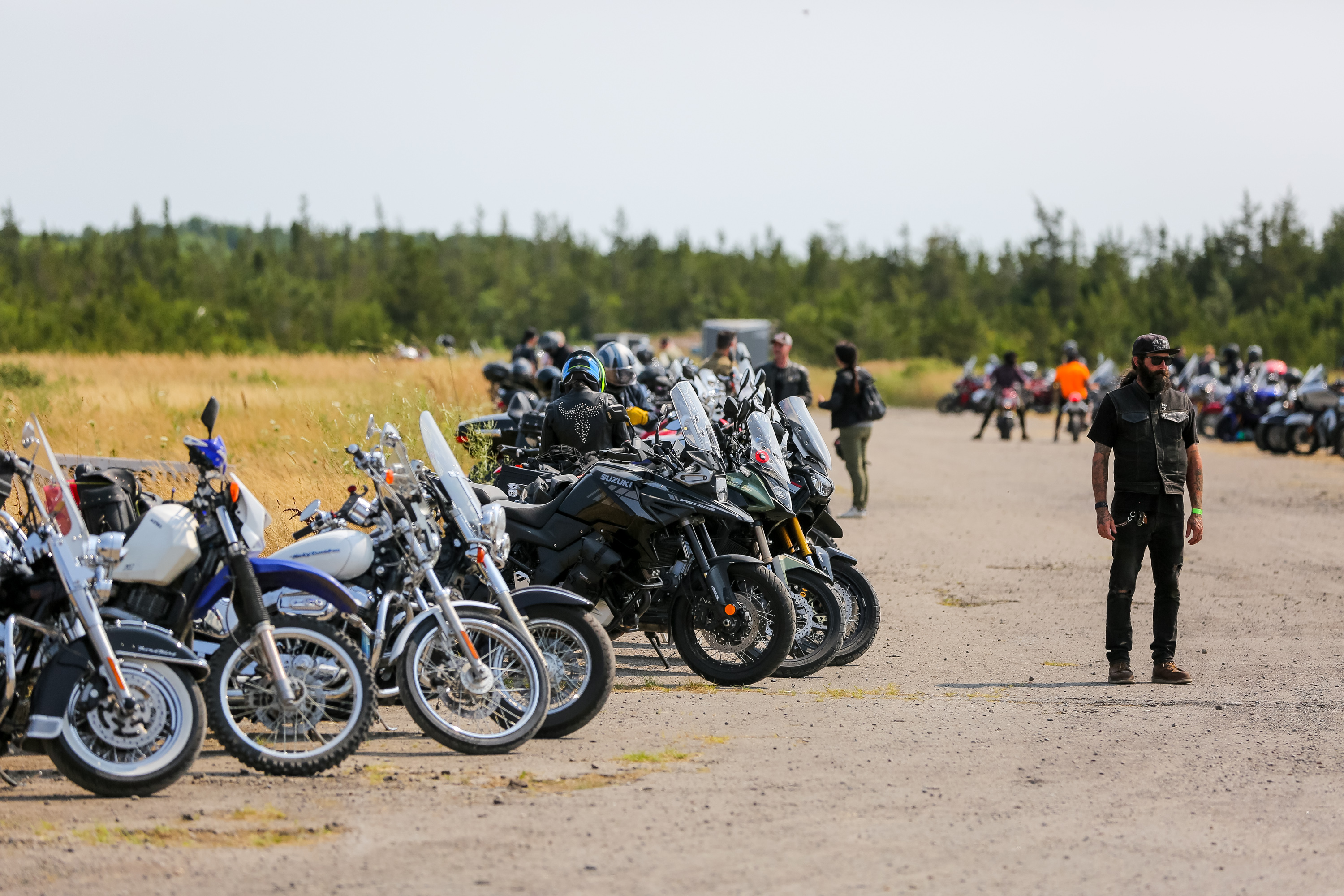 a rider stands in a parking lot filled with lines of motorcycles surrounded by green forest.