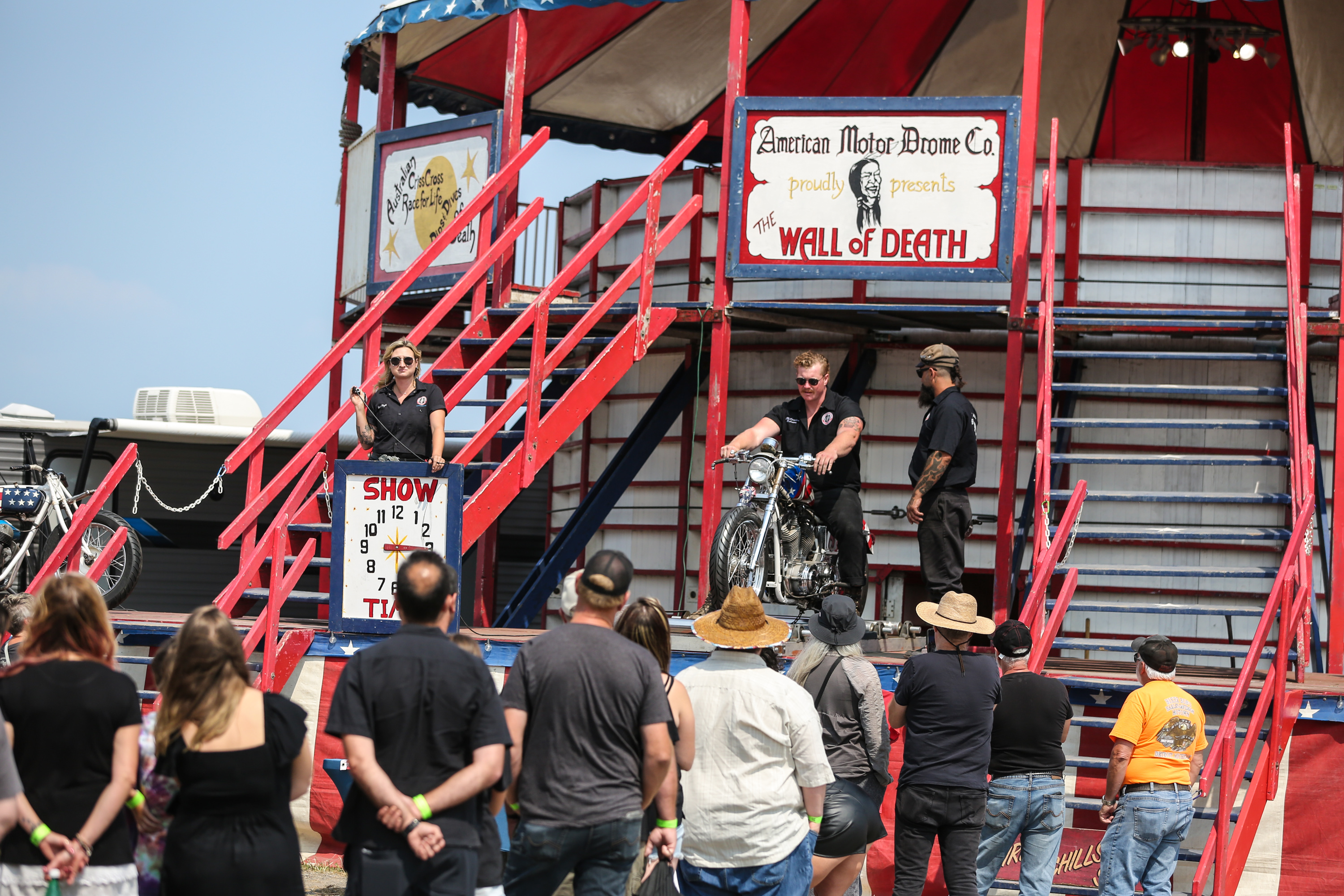 event patrons stand in line outside a tall wooden cylindrical structure, brightly coloured. A man on a motorcycle sits on a platform next to a female announcer, talking to the crowd under a vintage-inspired sign that reads "Wall of Death".