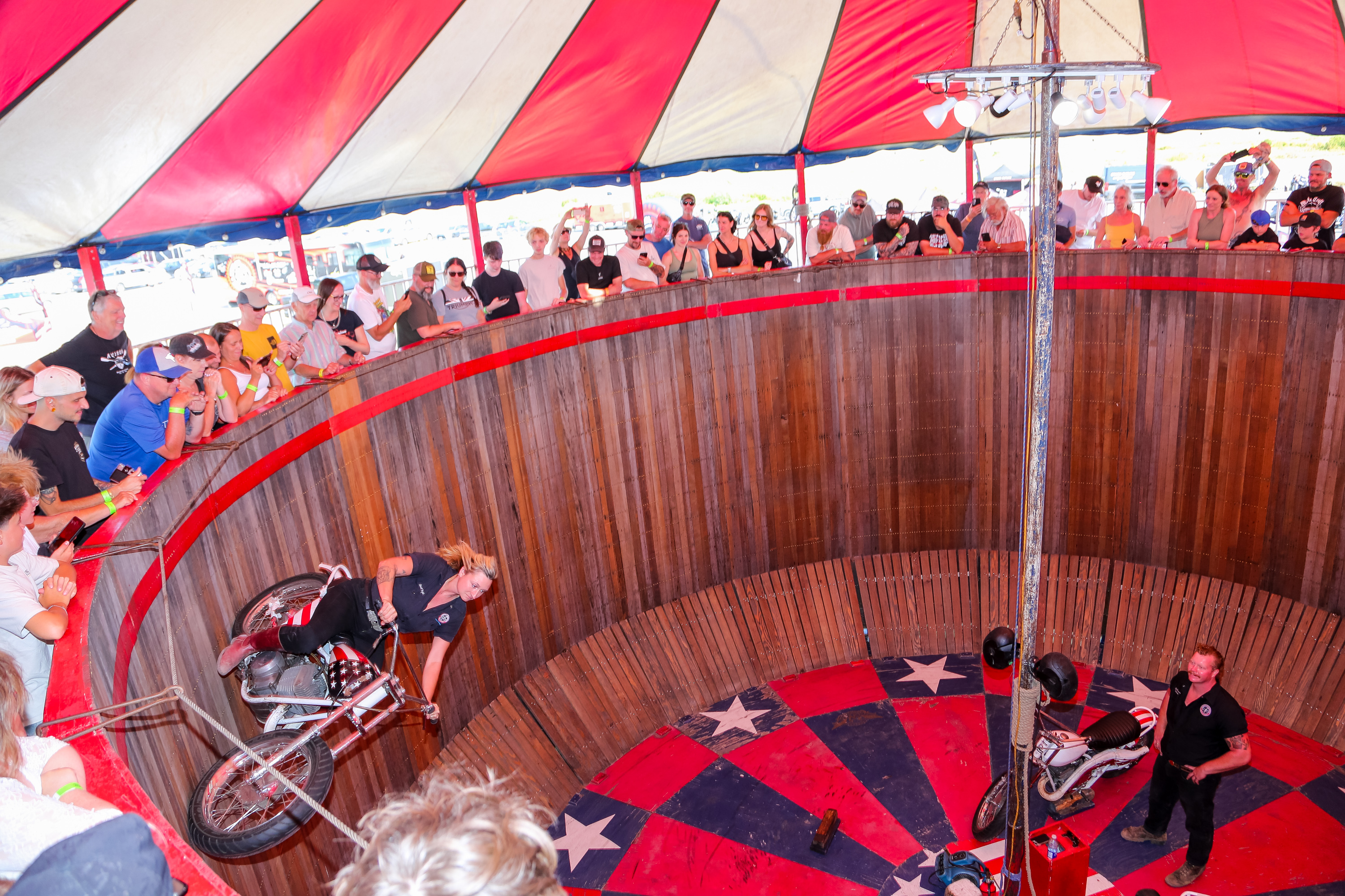 a female rider drives a motorcycle along the vertical, circular wooden wall of "The Wall of Death" at Moto Craft 2025, while a crowd looks on. 