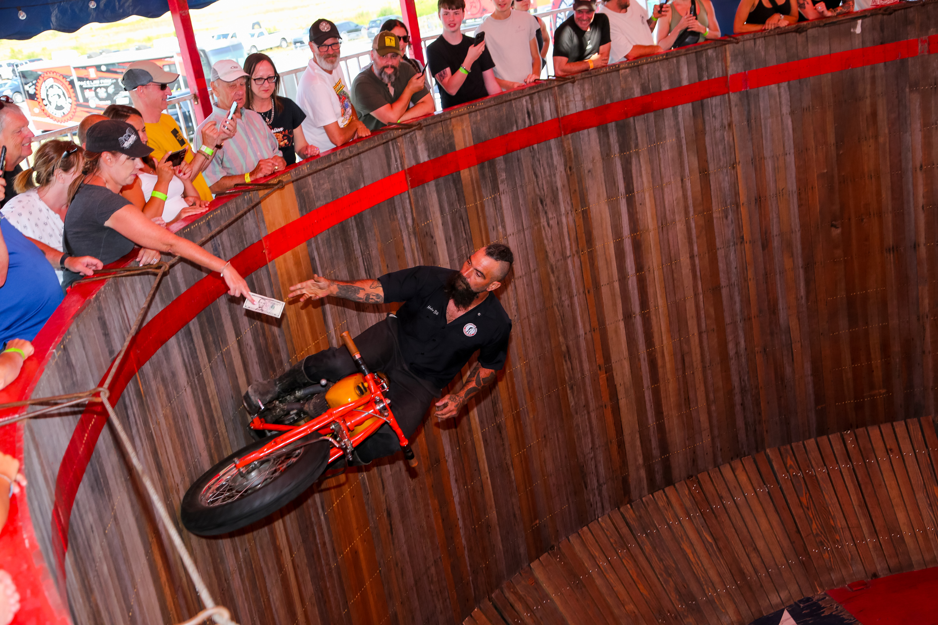 a man on a motorcycle rides along the vertical wooden wall of "The Wall of Death", collecting bills from the hands of members of the crowd at the top of the wall. 