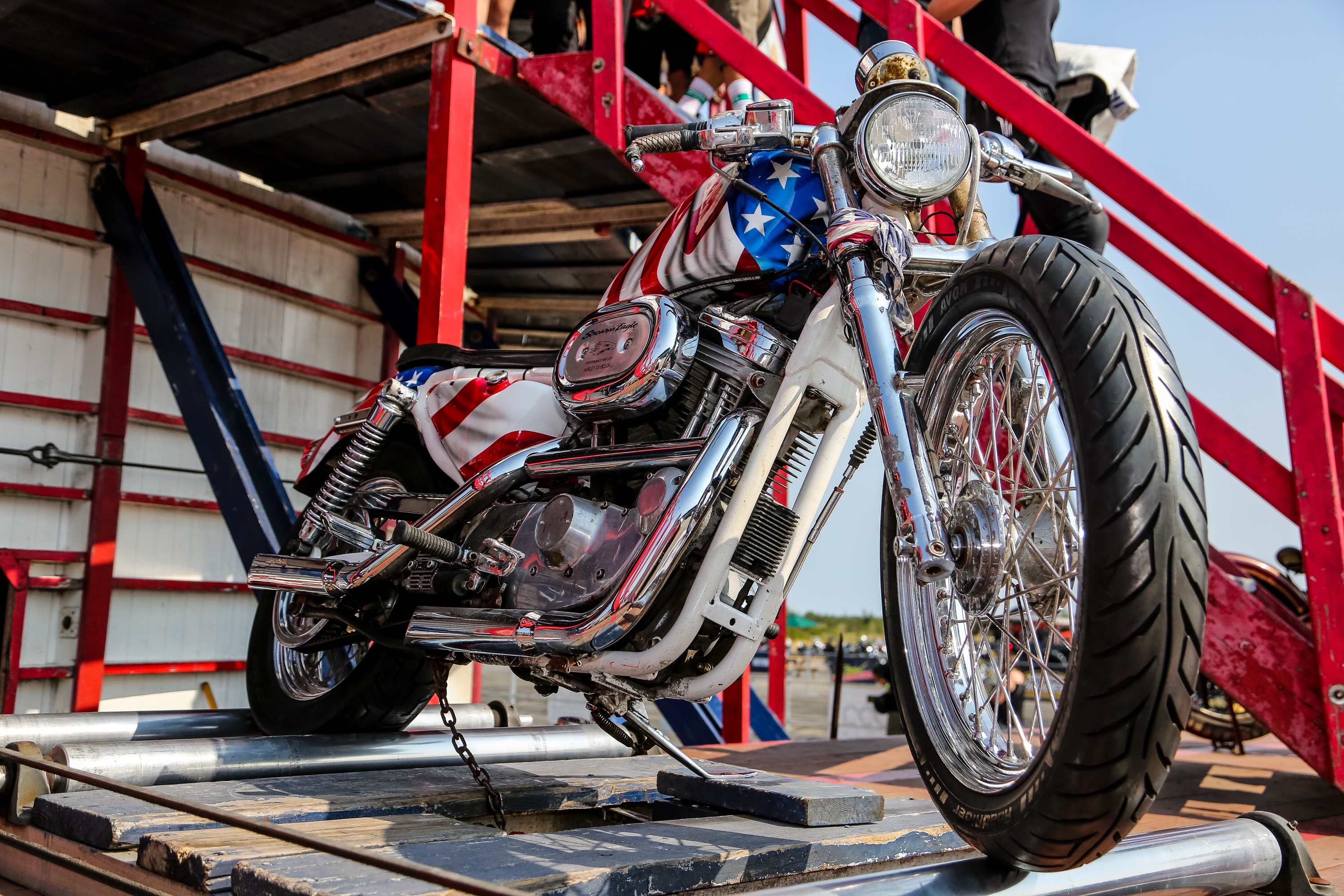 A shiny motorcycle painted with American Stars and Stripes sits on a platform next to the steep red wooden stairs to the "Wall of Death" attraction at Moto Craft on a sunny summer afternoon. 