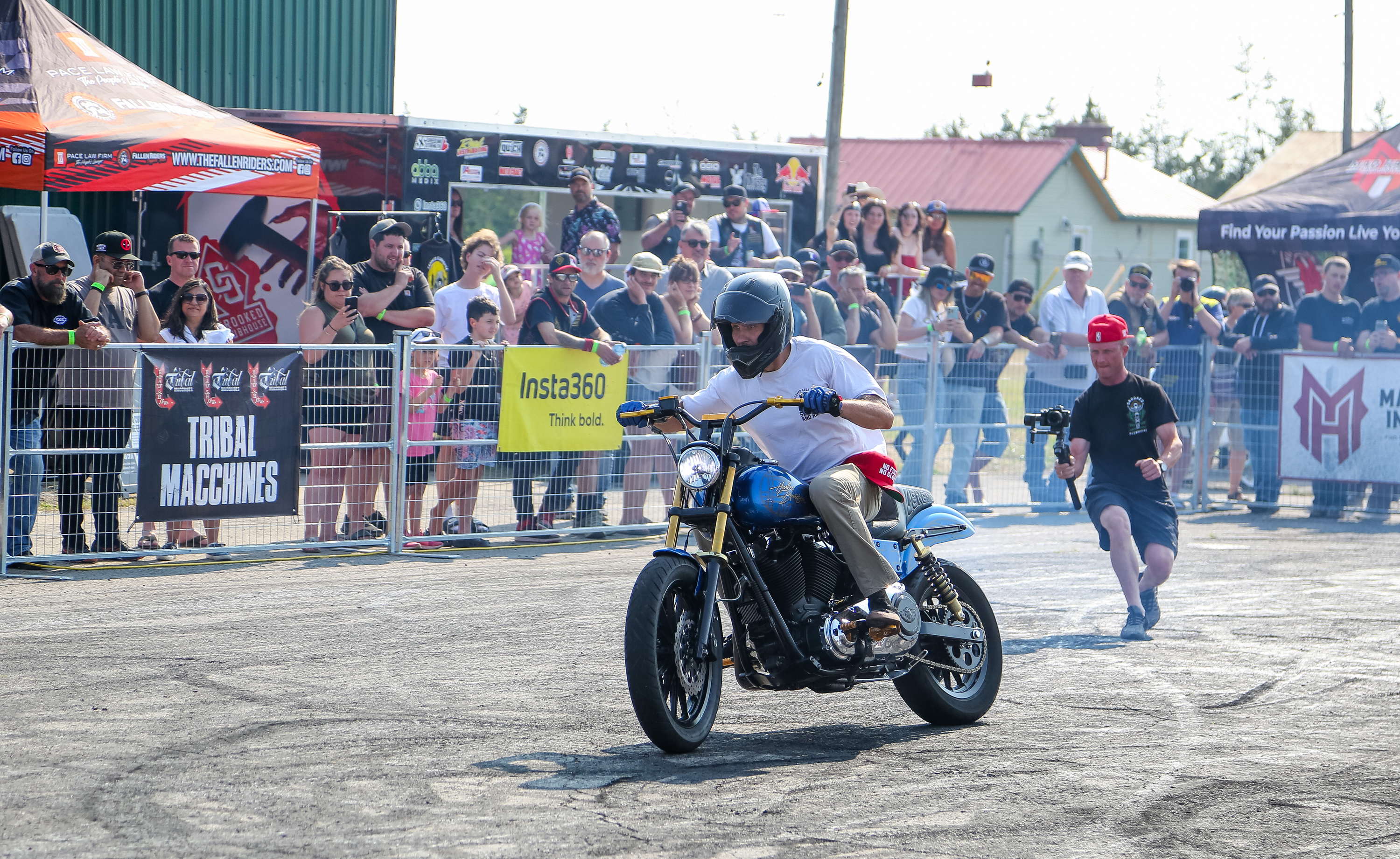 a rider races his motorcycle along a dirt stunt track in front of a large crowd, while a cameraman rushes behind him to catch the action.