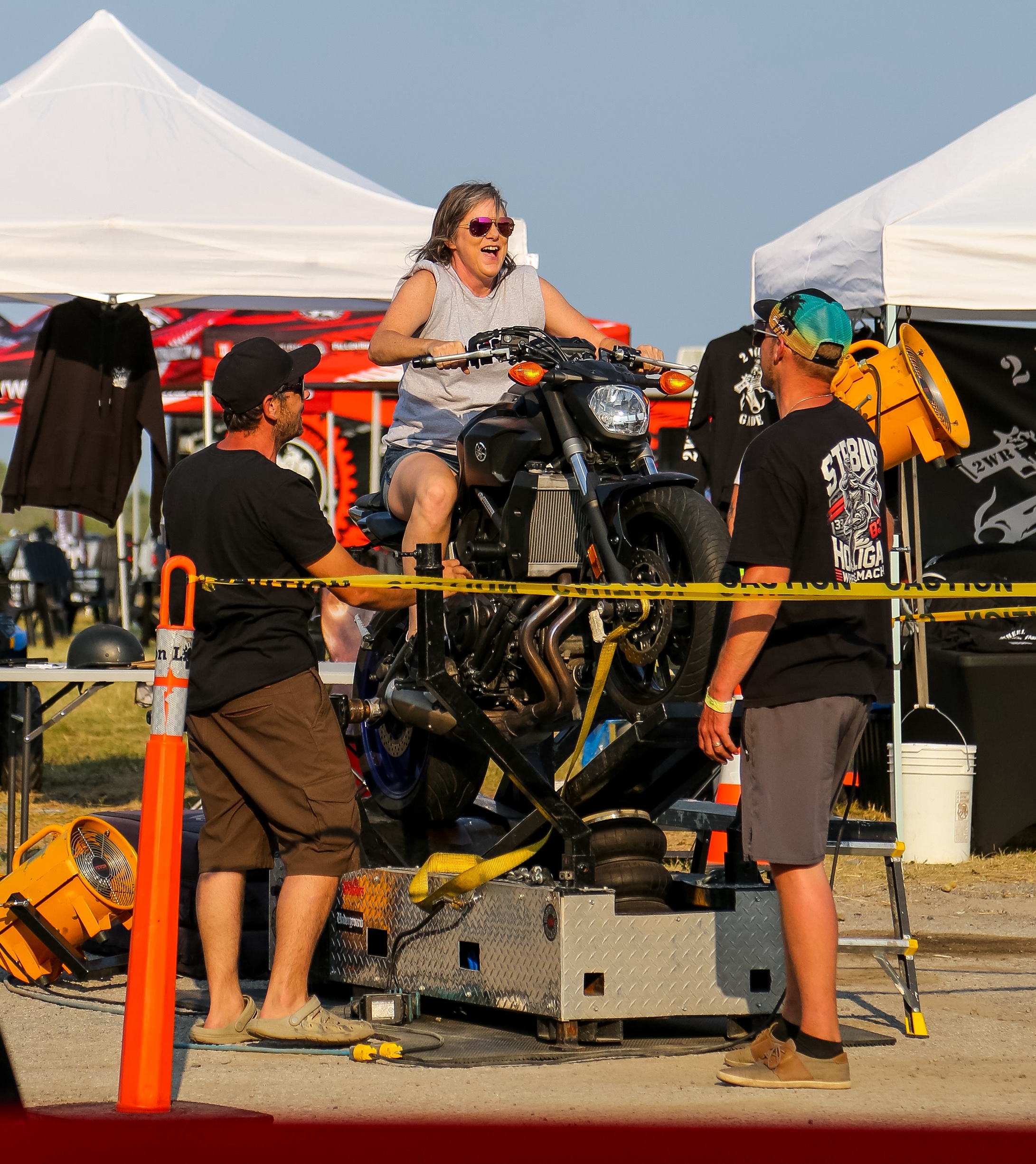 an older woman grins with excitement and chats to the staff assisting her while on the back of a motorcyle attached to a Wheelie Machine.