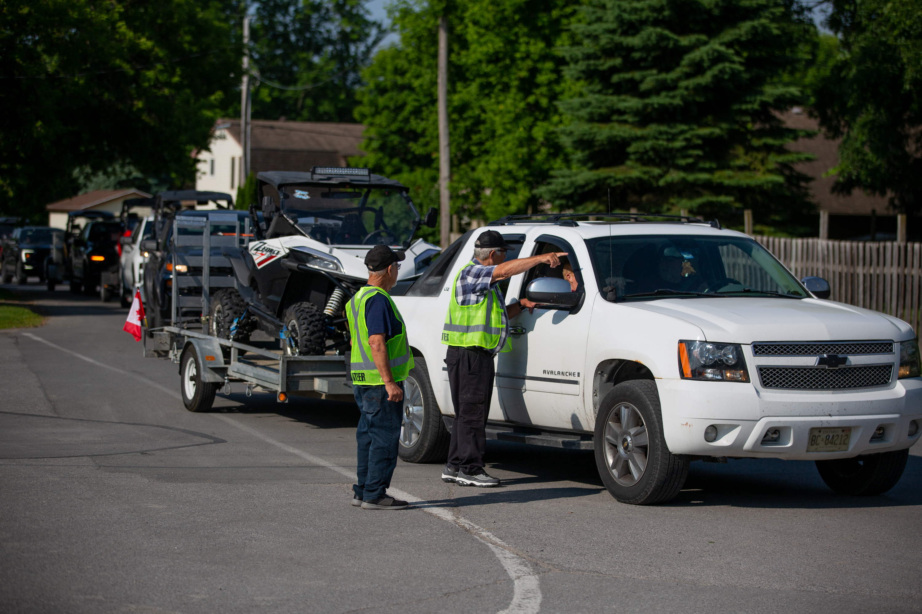 a long line of vehicles hauling ATVs stretch out behind two event staff in fluorescent green vests, who are talking to the driver of the lead vehicle and giving them directions.