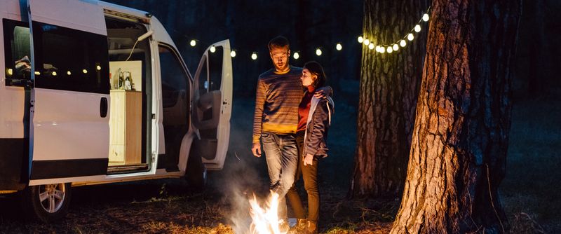 A couple stands with their arms around each other looking into a campfired next to their RV in the forest.