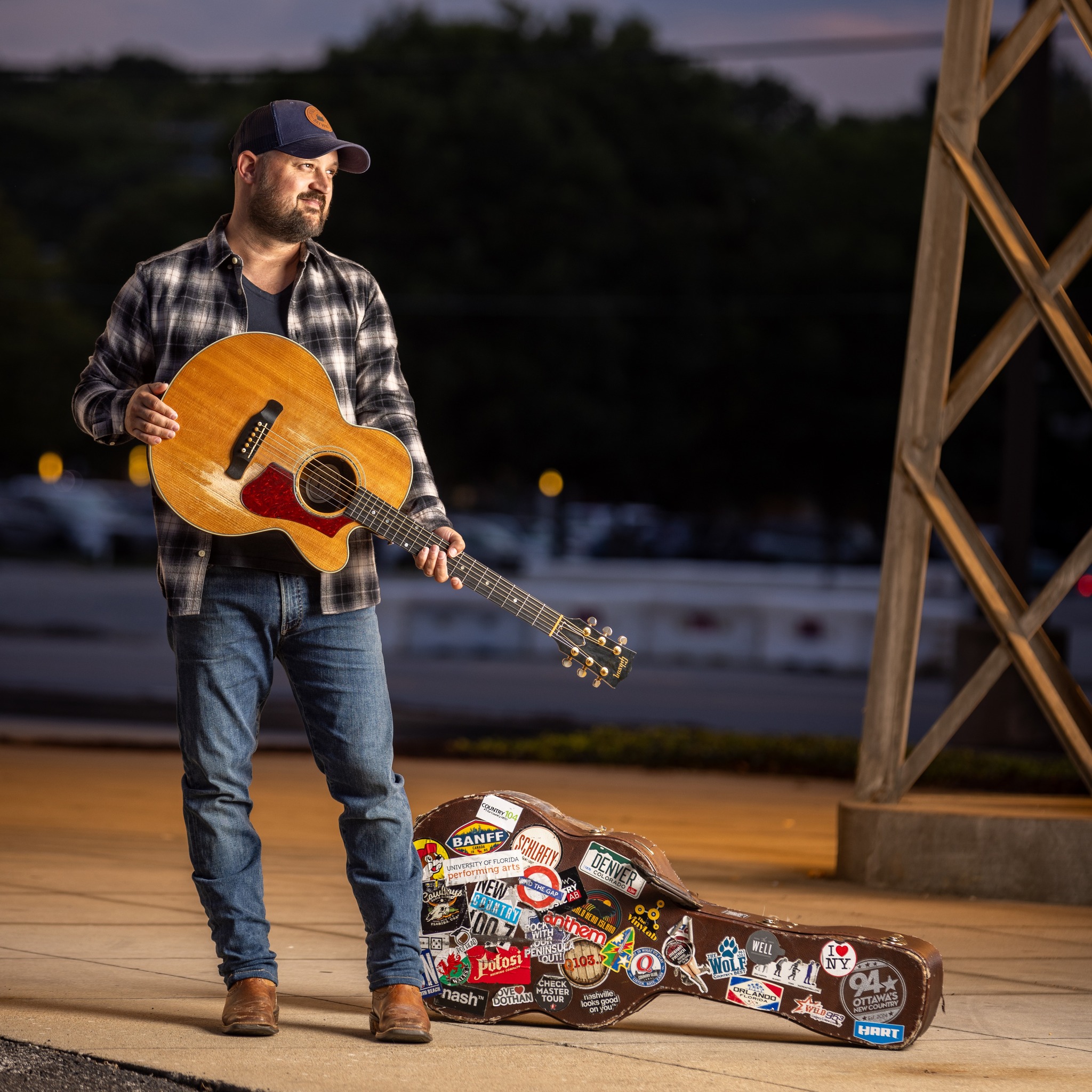 a smiling Aaron Goodvin standing outdoors under a spotlight and holding his guitar, next to a guitar case covered in travel stickers.