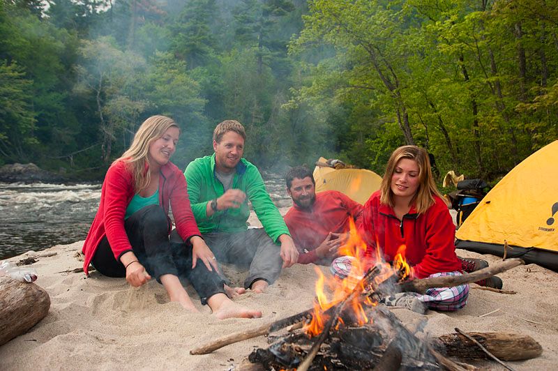 4 people talk and smile while gazing into their campfire on a soft sandy beach next to their tent at a river's edge surrounded by lush green forest.