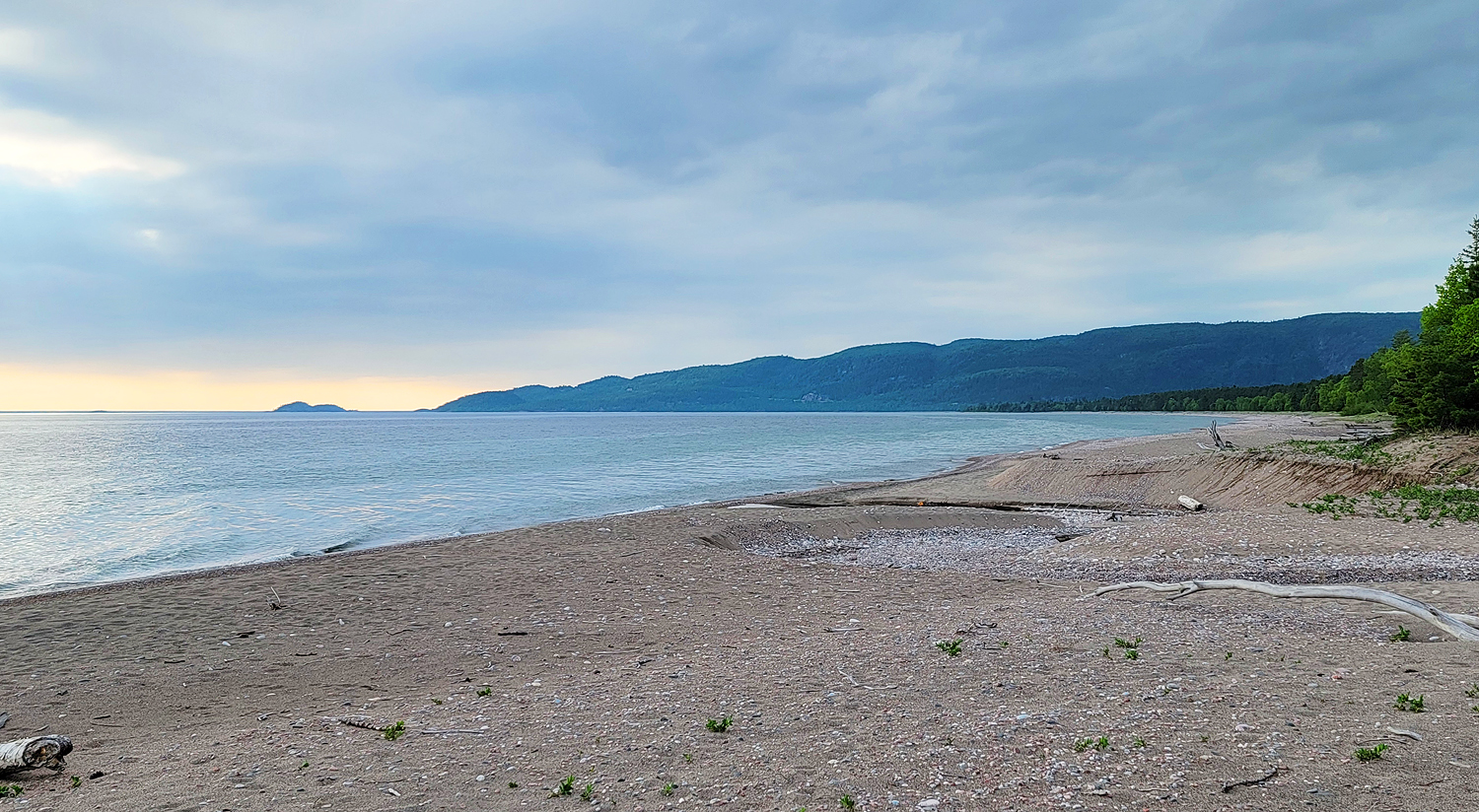 a grey pebble beach skirted by green boreal forest meets silvery grey Lake Superior waves under a breezy early evening sky.