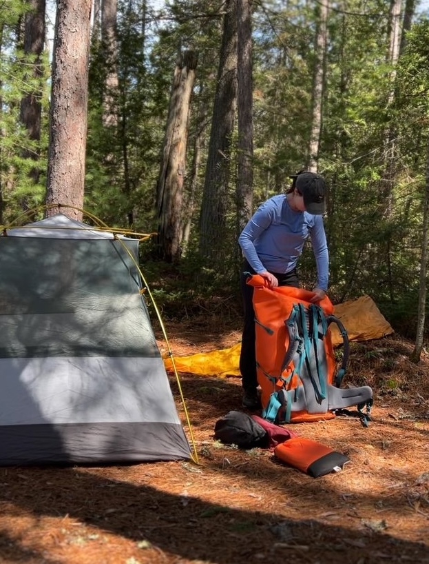 a camper rummages through their hiking pack next to a tent in a thick forest.