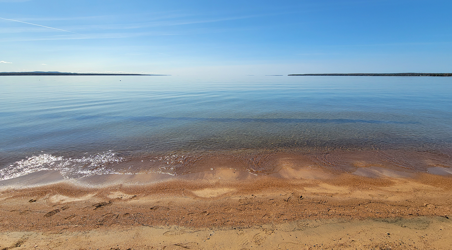 A golden sandy shore against an expanse of glassy blue water that meets the blue summer sky in an almost seamless transition.