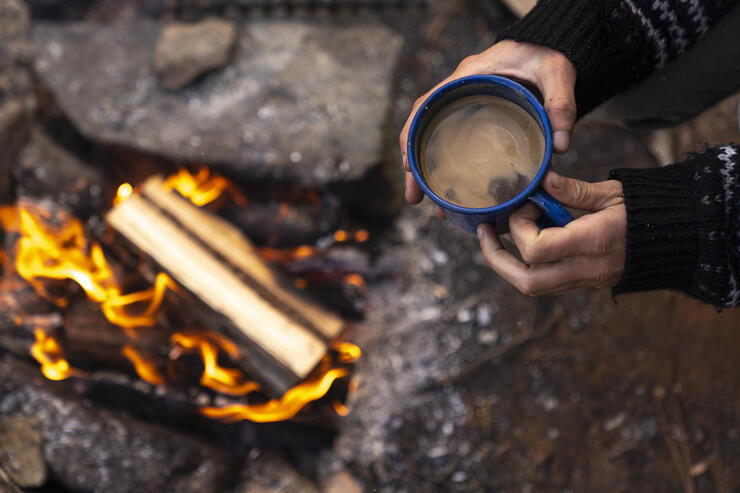 hands holding a steaming mug of camp coffee over a campfire surrounded by large grey stones